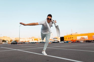 Modaspor ayakkabı güneş gözlüğü şık yazlık giysiler saç modeli ile genç Amerikalı hipster adam şehir otoparkında tek ayak üzerinde duruyor. Modern adam dengeyi korur. Gençlik sokak stili. Erkek giyim.