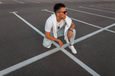 Şık bir vintage beyaz t-shirt bir sweatshirt ile şık kot moda genç hipster adam beyaz çizgileri ile bir yolda oturup. Kentsel modern adam sokakta rahatlatır. Erkek giyim.