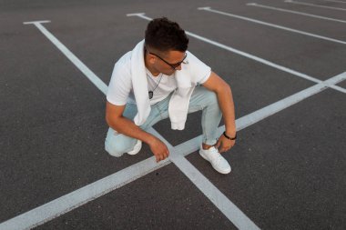 Şık bir saç modeli ile şık siyah güneş gözlüğü şık beyaz giysiler genç güzel adam hipster bir şehir otopark oturur. Yakışıklı Amerikalı erkek modeli açık havada rahatlatır. Yaz stili.