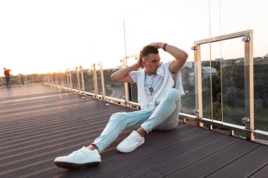 Terasta ahşap bir zemin üzerinde otururken poz beyaz spor ayakkabı vintage kot şık bir t-shirt şık bir saç modeli ile genç kentsel adam. Çekici adam modeli gün batımının tadını çıkarıyor. 