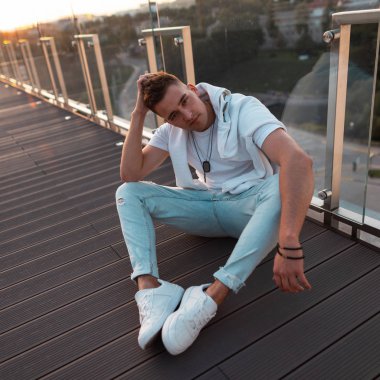 Şık yazlık giysiler modern güzel genç adam modeli gün batımında şehirde bir yaz açık terasta ahşap bir zemin üzerinde oturur. Çekici trendy adam açık havada dinlenme tadını çıkarıyor.