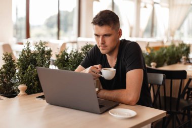 Siyah tişörtlü başarılı genç adam bir kafede bir fincan kahve ile oturur ve bir dizüstü bilgisayarda çalışır. Çekici serbest adam uzaktan çalışır. Çalışma süresi.