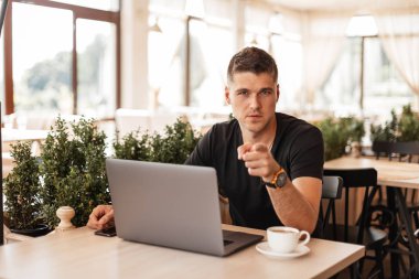 Yakışıklı genç iş adamı kahve ile bir dizüstü bilgisayar ile bir kafede oturuyor ve size parmağını işaret ediyor. Başarılı serbest adam uzaktan kapalı çalışan.