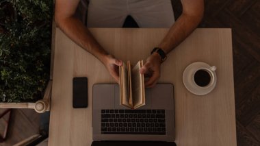 Genç başarılı adam bir cep telefonu ile kahve bir fincan ile modern bir dizüstü bilgisayar ile bir masada oturur ve elinde bir kitap tutar. Adam eski bir kafede oturan bir kitap okuyor. Üst te. Close.