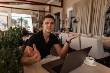 Şık bir t-shirt yakışıklı genç iş adamı bir kafede bir fincan kahve ile modern bir dizüstü bilgisayar ile sevimli bir gülümseme ile çalışır. Başarılı pozitif iş adamı uzaktan kapalı çalışan. Çalışma süresi.