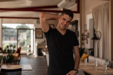 Bir yaz güneşli bir günde bir vintage kafede ayakta poz şık bir siyah t-shirt şık bir saç modeli ile güzel bir gülümseme ile Pozitif genç adam modeli. Yakışıklı neşeli adam dinlenme tadını çıkarıyor.