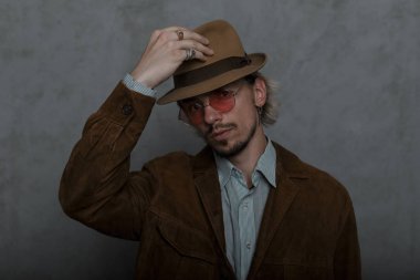 Klasik bir gömlek ayakta kahverengi moda ceket yuvarlak vintage gözlük bir sakallı eski moda genç adam hipster ve gri bir duvarın yakınındaki bir odada şık bir şapka doğrultar. Yakışıklı adam. 