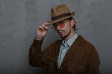 Şık genç sakallı hipster adam pembe yuvarlak gözlük şık bir şapka içinde şık bir kahverengi ceket klasik bir gömlek stüdyoda gri bir duvar yakınında poz. Çekici erkek modeli. Retro tarzı.