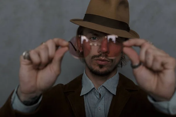 Portret van een stijlvolle jonge hipster man met een baard in een luxe hoed in een bruin Vintage jasje in een shirt. Knappe kerel shows in de camera trendy rode ronde glazen. Close-up. Focus op bril. — Stockfoto