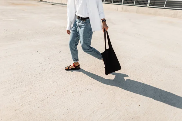 Closeup of male legs in stylish jeans in red leather summer shoes with a cloth black vintage bag. Young urban man goes on the pavement. New trendy collection of men\'s shoes. Street style.