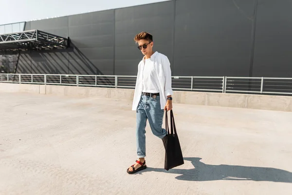 Moderní mladý muž hipče v módní bílé a Džínové Oblečení v slunečních brýlích v červených sandálech a ve slunném dnu chodí na ulici s pytlíčkem. Městský chlápek na dovolené. Pánské stylové šaty. — Stock fotografie