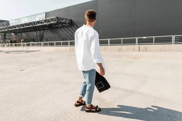 Πίσω όψη σε ένα μοντέρνο νεαρό άντρα σε κομψά ρούχα σε ένα κόκκινο καλοκαίρι παπούτσια με ένα μοντέρνο χτένισμα με ένα πανί μαύρη σακούλα. Σύγχρονος τύπος περπατάει γύρω από την πόλη σε μια φωτεινή ηλιόλουστη μέρα. Στυλ δρόμου. — Φωτογραφία Αρχείου