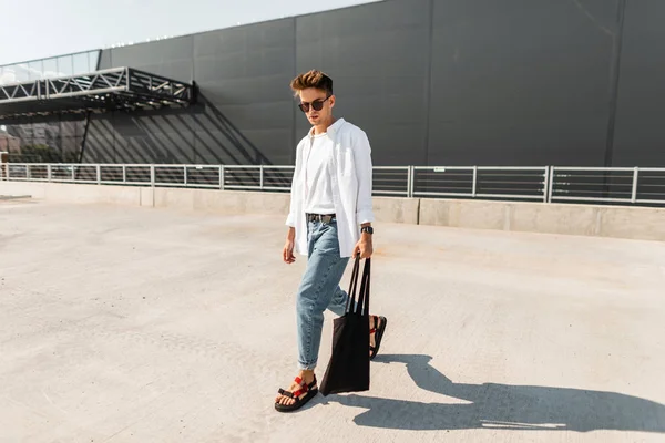 Amerykański model mody młodego mężczyzny w modnych dżinsów w stylowej koszulce w modne sandały w okulary przeciwsłoneczne z torbą spacery w pobliżu szarego budynku. Nowoczesny facet Hipster. Nowa letnia kolekcja odzieży męskiej. — Zdjęcie stockowe