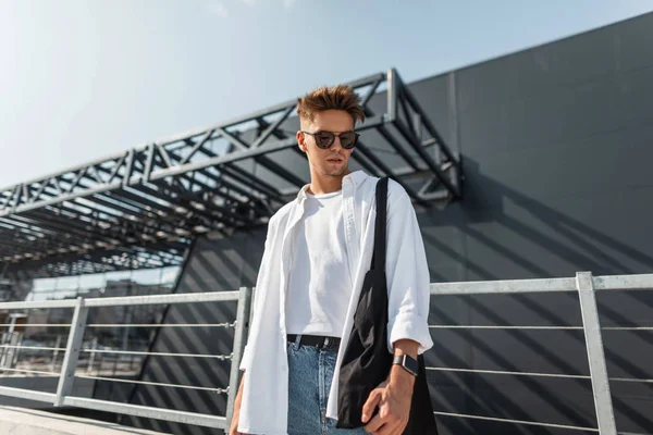 Stijlvolle jonge man hipster in donkere zonnebrillen in witte modieuze kleding met kapsel met zwarte tas staat in de buurt van een grijs gebouw op een zonnige dag buitenshuis. Trendy kerel model. Zomer jeugd stijl. — Stockfoto