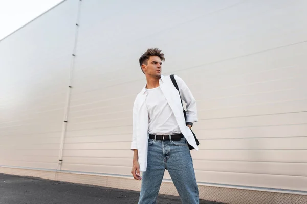 Knappe jonge man in blauwe jeans met een stijlvolle kapsel in modieuze shirt met stof Vintage tas in de buurt van een wit gebouw op een zomerse dag buitenshuis. Amerikaanse kerel. Nieuwe zomer collectie herenkleding. — Stockfoto