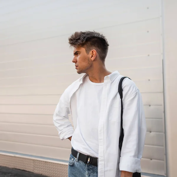 Young attractive man model in an elegant white shirt in a T-shirt in blue stylish jeans with a vintage bag stands on the street near a white modern wall. American guy. Youth street style.