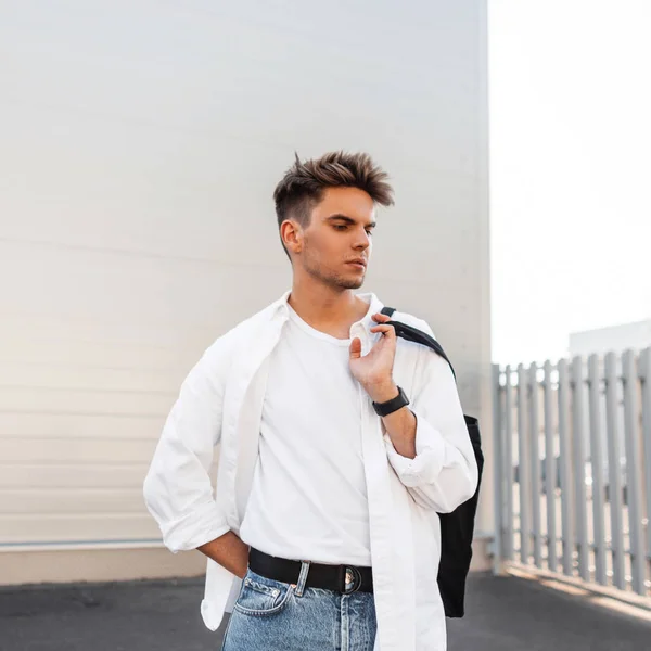 Aantrekkelijke stijlvolle jonge man in een elegant wit overhemd in blauwe jeans met een modieuze tas met een trendy kapsel poseren in de buurt van een wit gebouw in de stad. Knap stedelijk model Guy buitenshuis. — Stockfoto