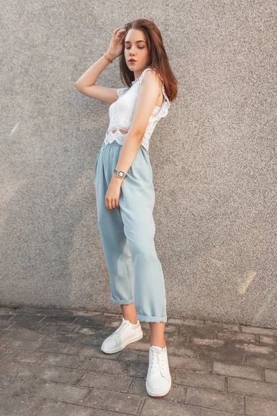 Mulher jovem e elegante urbana em um top de renda em calças azuis em tênis posando perto de parede cinza na rua. Modelo menina muito atraente gosta da luz do sol na cidade em um dia de verão . — Fotografia de Stock