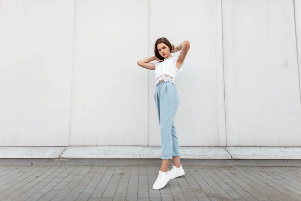 Elegantní, krásná mladá žena v bílé, módní krajkové blůze ve stylových teniskách v módních modrých kalhotách, které se nacházejí nedaleko starobylé zdi ve městě. Evropská dívčí modelka na procházce za letního dne. — Stock fotografie