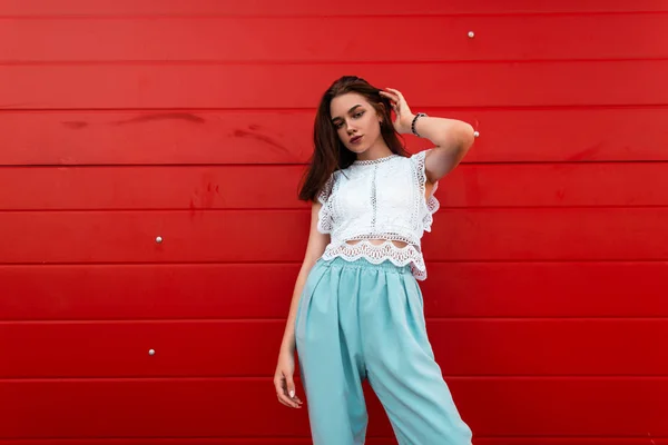 Stedelijke moderne jonge vrouw recht haar. Vrij sexy meisje in een modieuze witte Lace blouse in blauwe broek poseren op de straat in de buurt van een rode muur. Trendy zomer collectie Dameskleding. — Stockfoto