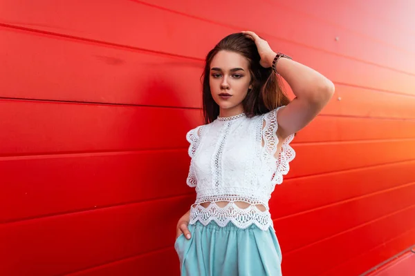 赤い木製の壁の近くの街でポーズヴィンテージブルースパンツでスタイリッシュなレースの白いブラウスでかなりエレガントな若い女性モデル。現代のかわいい女の子のブルネット屋外。スタイリッシュな女性の夏の服. — ストック写真