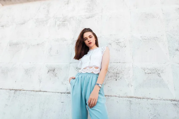 Hübsche junge Frau Modell in Vintage-Spitze weiße Bluse in trendigen blauen Hosen posiert in der Stadt in der Nähe der weißen Wand. attraktive elegante Brünette Mädchen im Freien. stilvolle Sommerkleidung für Frauen. — Stockfoto