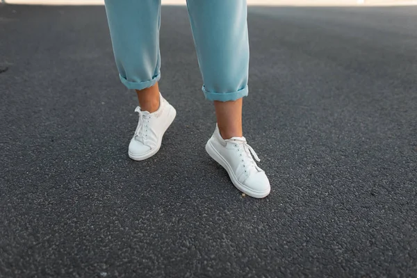 Vrouwelijke benen in stijlvolle blauwe broek in wit lederen sneakers op het asfalt in de stad. Trendy jonge vrouw op een wandeling. Modieuze Damesschoenen. Nieuwe collectie. — Stockfoto