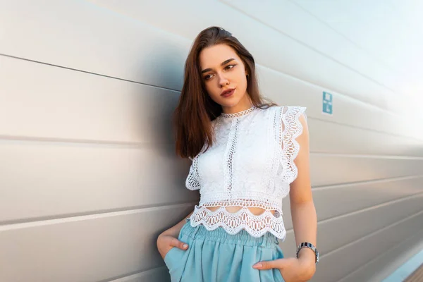 Moderní, velmi mladá modelová žena ve stylové krajkové blůze v módních modrých kalhotách, které se nacházejí u bílé zdi ve městě. Moderní elegantní dívka odpočívá venku. Letní módní vzhled. — Stock fotografie