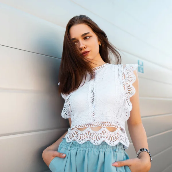 Ung elegant kvinna mode modell i en fashionabel spetstopp i snygga blå byxor står i staden nära en vit Vintage Wall. Modern trendig tjej vilar på gatan. Sommar stil. — Stockfoto