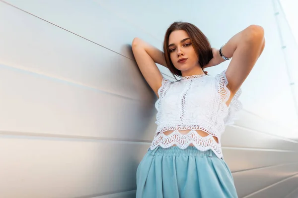 Stylische junge brünette Frau in modischen weißen Spitzenbluse in Vintage-blauen Hosen ruht draußen in der Nähe einer modernen Wand. ziemlich elegantes Mädchen Modell entspannt sich im Freien an einem Sommertag. neue Kollektion. — Stockfoto