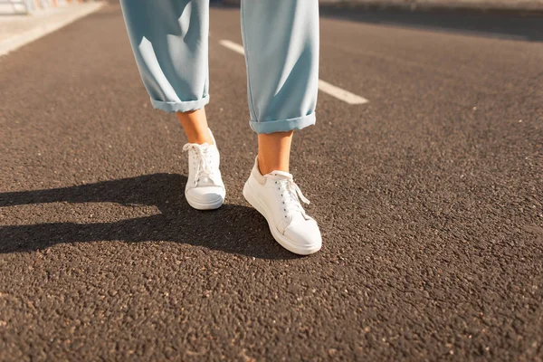 Close-up modieuze vrouwelijke benen in stijlvolle blauwe broek in een witte trendy sneakers staan op het asfalt op een heldere zonnige dag. Modieuze moderne zomer collectie van damesschoenen. Casual stijl. — Stockfoto