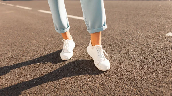 Närbild av kvinnliga ben i blå Vintage byxor i snygga lädersneakers på asfalten. Fashionabla kvinna på en promenad på en solig ljus sommardag. Modern säsongsbetonad kollektion av snygga sneakers. — Stockfoto