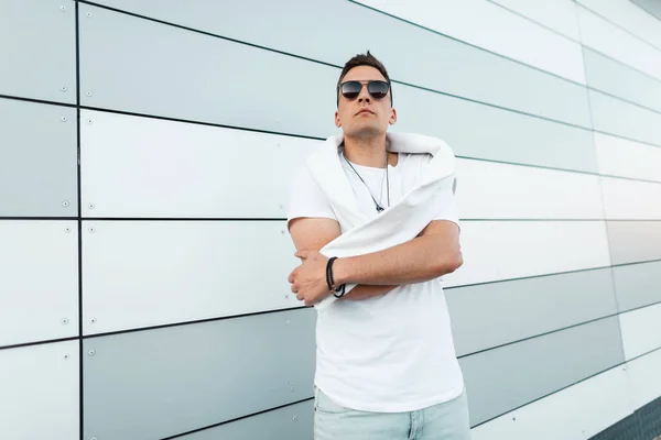 Modelo de moda de hombre joven con sudadera blanca en una elegante camiseta con gafas de sol negras de moda posando cerca de una pared moderna en la ciudad. Americano guapo hipster chico descansando al aire libre . — Foto de Stock