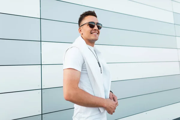 Alegre joven hipster hombre en gafas de sol en una camiseta blanca de moda con un medallón en el cuello se encuentra cerca de un moderno edificio a rayas en la ciudad en un día de verano. Feliz modelo de buen tipo. Moda callejera — Foto de Stock