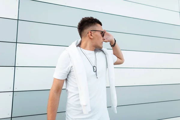 Urban Handsome Young hipster man i en trendig vit t-shirt i snygga solglasögon med fashionabla frisyr poserar nära en modern byggnad i staden. Attraktiv kille modell utomhus. Sommar stil. — Stockfoto