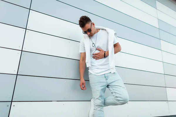 Trendy young hipster man in a fashionable white t-shirt in dark sunglasses in ripped jeans posing near a modern building on the street. Handsome guy model outdoors. Summer youth fashion.Trendy clothes — Stock Photo, Image