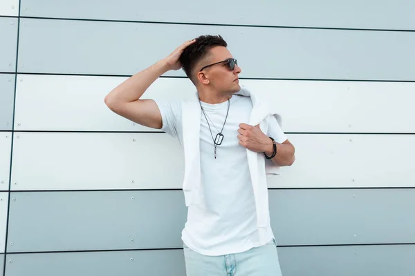 El modelo a la moda de hombre joven en la ropa de verano de estilo en gafas de sol de moda posando en la ciudad cerca de una pared moderna. Un americano urbano hipster al aire libre. Moda juvenil callejera. Prendas masculinas . — Foto de Stock