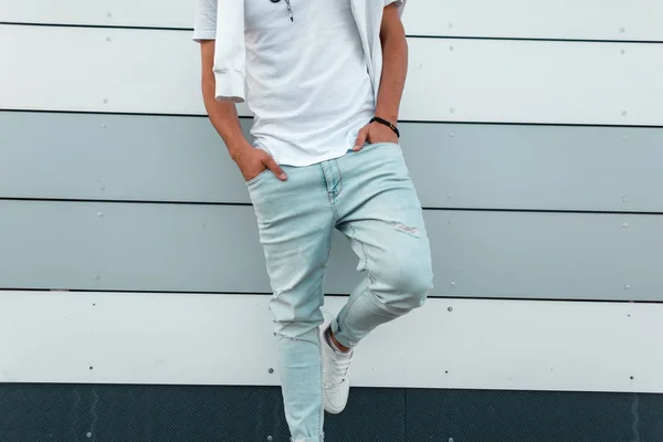 Fashionabla ung man i blå trendiga jeans i en snygg vit t-shirt i läderskor poserar nära en modern byggnad i staden. Ny kollektion av herrsommarkläder. Ungdoms stil. Närbild. — Stockfoto