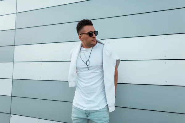 Coole junge amerikanische Hipster mit Sonnenbrille in blauen trendigen Jeans in einem modischen weißen T-Shirt posieren in der Stadt in der Nähe einer modern gestreiften Wand. netter Kerl mit Frisur im Freien an einem Sommertag. — Stockfoto