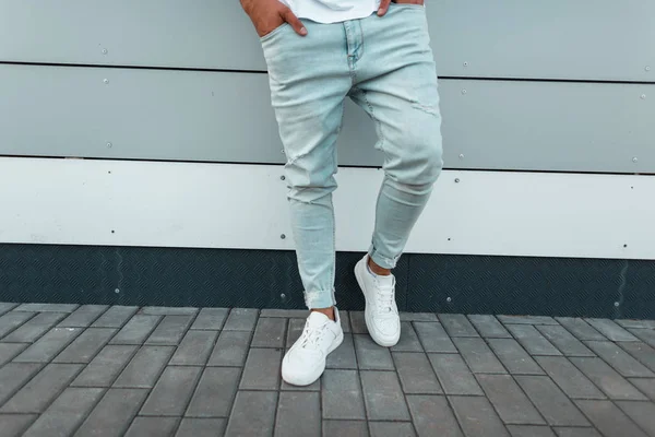 Μοντέρνο νεαρό άνδρα σε ένα t-shirt σε μοντέρνα άρπαξαν μπλε τζιν σε κομψά λευκά δερμάτινα αθλητικά παπούτσια στέκεται στο δρόμο κοντά στον τοίχο. Σύγχρονος τύπος στην πόλη. Καλοκαιρινά αντρικά ρούχα. Γκρο πλαν. — Φωτογραφία Αρχείου