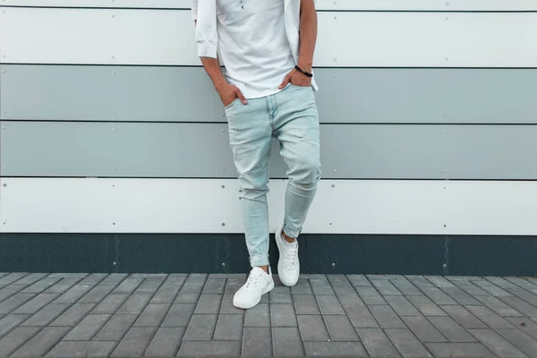 Νέος κομψός άνθρωπος σε ένα λευκό μοδάτο μπλουζάκι σε vintage μπλε τζιν σε λευκά μοντέρνα αθλητικά παπούτσια στέκεται κοντά σε ένα σύγχρονο τοίχο. Μοντέρνα ανδρικά καλοκαιρινά ρούχα. Το στυλ του δρόμου. Κοντινό πλαίσιο του αρσενικού σώματος. — Φωτογραφία Αρχείου
