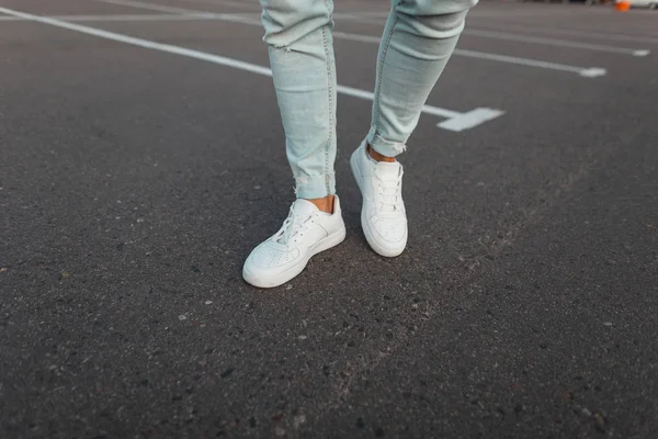 Närbild på manliga ben. Trendiga unga fashionabla mannen i Vintage blå jeans i läder snygga vita sneakers står på asfaltvägen utomhus. Modern ungdoms gata stil. — Stockfoto
