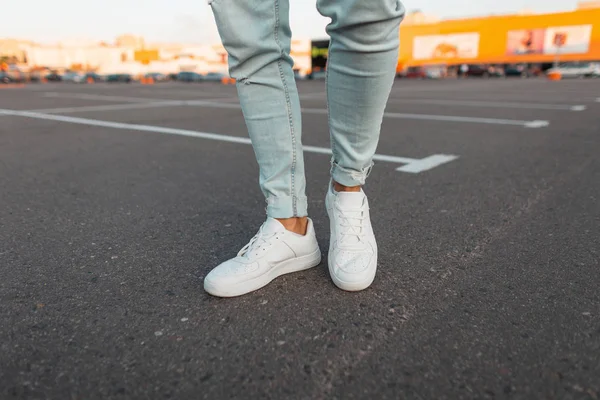 Modieuze moderne jonge man in stijlvolle blauwe jeans in een trendy leren seizoensgebonden witte sneakers staat op een parkeerplaats in de stad. Close-up van mannen stijlvolle benen. Zomerlook. — Stockfoto