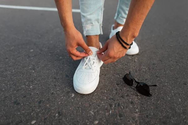 Jonge stijlvolle man in blauwe jeans in lederen witte trendy schoenen staat op de weg naast zonnebril en rechtzetten schoenveters. Modieuze heren sneakers en accessoires. Close-up. — Stockfoto