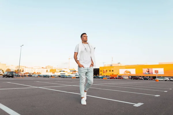 Knappe trendy jonge man in modieuze blauwe jeans in een stijlvol wit T-shirt in Sneakers wandelingen rond de stad op een heldere zomerdag. Moderne kerel model rusten op de straat. Zomer Streetstyle. — Stockfoto
