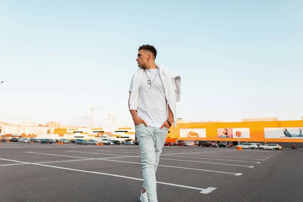 Μοντέρνο νεαρό άντρα με ένα κομψό χτένισμα σε μπλε τζιν σε ένα λευκό μπλουζάκι με ένα πουλόβερ είναι περπατώντας στο χώρο στάθμευσης στην πόλη. Ο Αστικός σύγχρονος τύπος απολαμβάνει το καλοκαιρινό Σαββατοκύριακο. Στυλ νεολαίας του δρόμου — Φωτογραφία Αρχείου