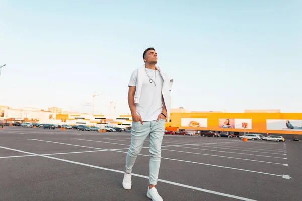 Jonge knappe man model in modieuze zomer wit en denim kleding in lederen sneakers met een kapsel wandelingen in een stad parkeren. Stijlvolle Amerikaanse kerel geniet van een wandeling. Modieuze zomer herenkleding. — Stockfoto