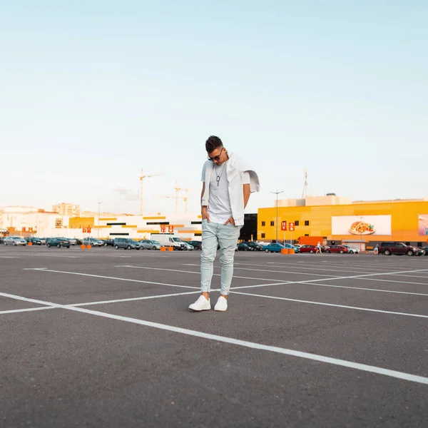 Beyaz spor ayakkabılı, şık tişörtlü, tişörtlü, mavi kot pantolonlu güneş gözlüklü Amerikalı genç hipster adam caddeden aşağı iner. Otoparkta yakışıklı bir şehirli. Şık yaz erkek giyim.