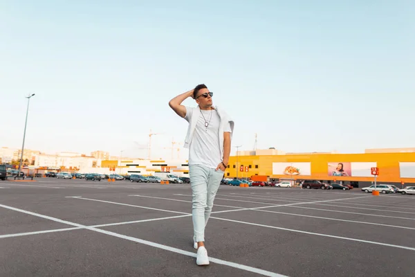 Stijlvolle stedelijke jonge man in zonnebril in modieuze zomer kleding in leer witte trendy sneakers geniet van een wandeling door de stad. Trendy kerel hipster met stijlvolle kapsel in een parkeerplaats buiten. — Stockfoto