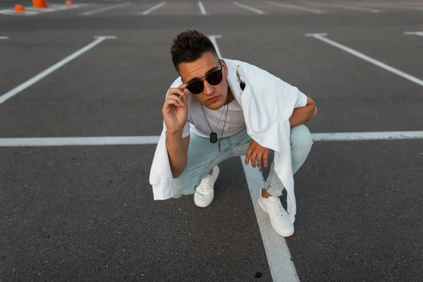 Bir asfalt üzerinde oturan bir sweatshirt ile bir moda t-shirt beyaz spor ayakkabı lı mavi kot pantolon modern genç hipster adam ve kamera bakmak. Serin şehir adam otoparkta rahatlatır. Şık erkek giyim.